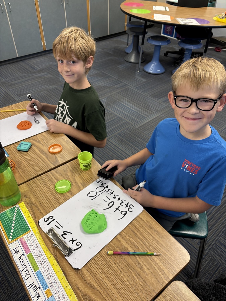 2 students sitting a table with whiteboards present and play dough visible. Students have multiplication problems on white boards.