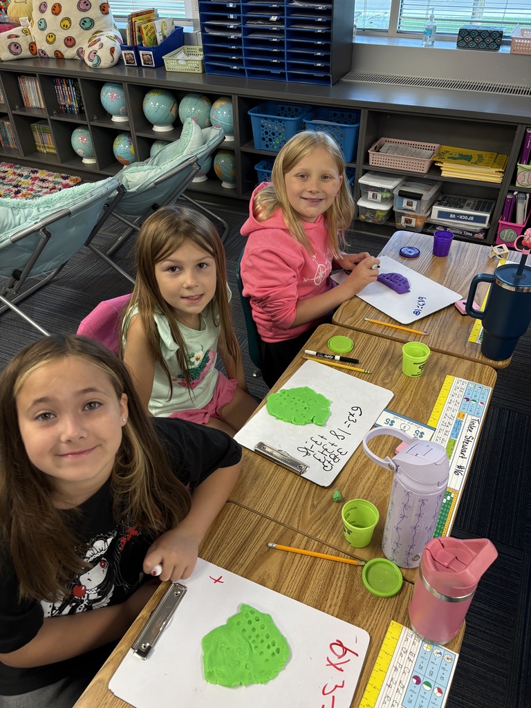 3 students sitting a table with whiteboards present and play dough visible. Students have multiplication problems on white boards.