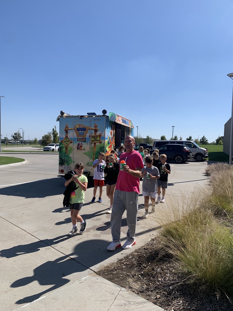 	A man in a bright pink shirt and a group of children walk away from the Kona Ice truck (visible in the background), all holding their cups of shaved ice on a sunny day.