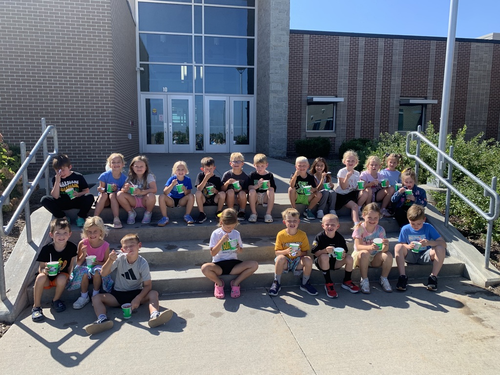 A large group of elementary-aged children is sitting on the steps outside a school entrance, all holding their cups of shaved ice and smiling for the camera.