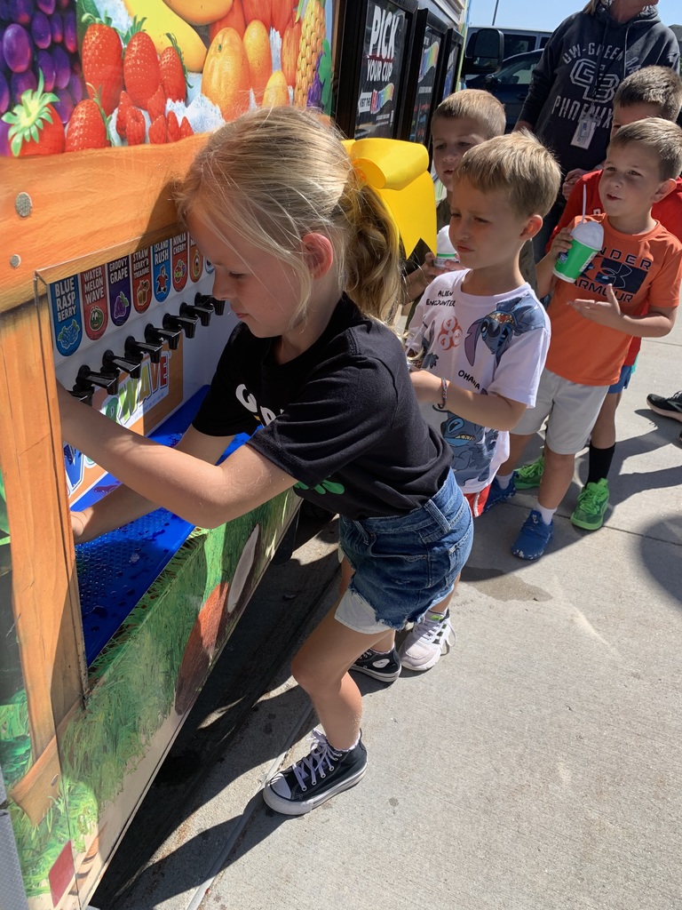 A young blonde girl in a black shirt and denim shorts is focused on dispensing flavor onto her shaved ice from the Kona Ice Flavorwave, as three young boys wait in line behind her.