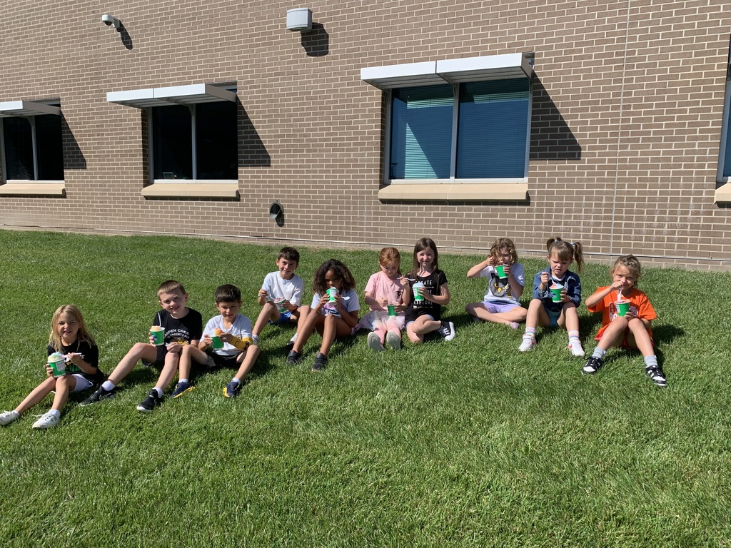 	A group of nine young children are sitting in a row on a sunny patch of green grass outside a brick building, happily eating their cups of shaved ice.