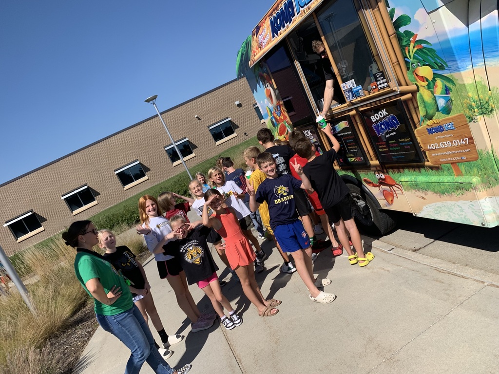 A group of laughing children are gathered around the side of the Kona Ice truck, with some lining up to receive their cups. A woman in a green shirt watches them on the left.