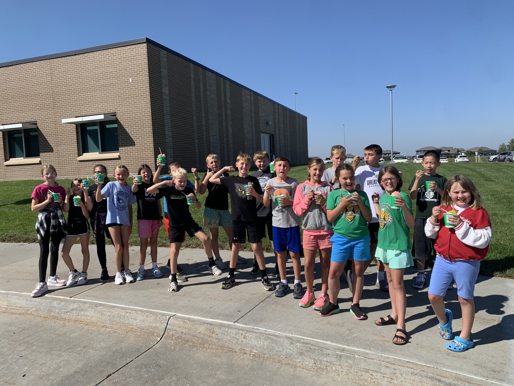 A line of children stands outside a brick school building on a sunny day, holding their shaved ice cups and posing with big smiles for the picture.