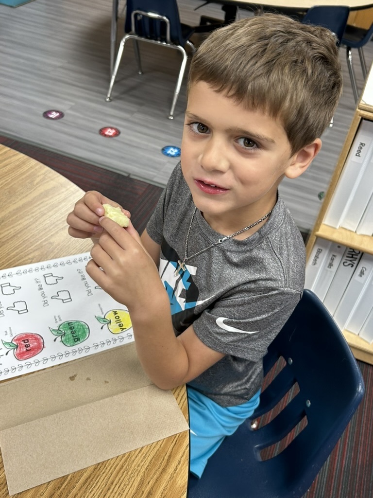 Boy sitting at a table smiling while eating an apple in front of a paper that shows apples.