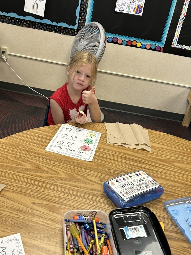 Girl sitting at a table with an apple in her hand with her thumb up and a paper on the table that says Apple Taste Test.