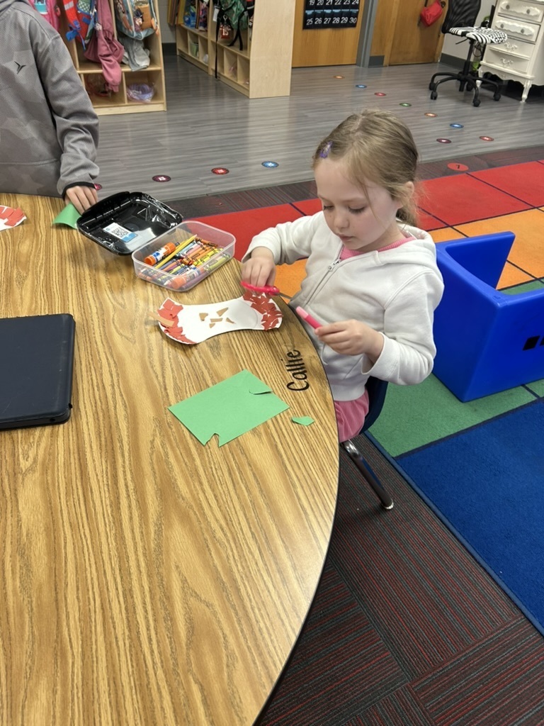 Girl sitting in a chair working at a table on a craft that looks like an apple.