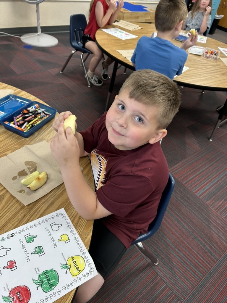 Boy sitting at a table smiling while eating an apple in front of a paper that shows apples.