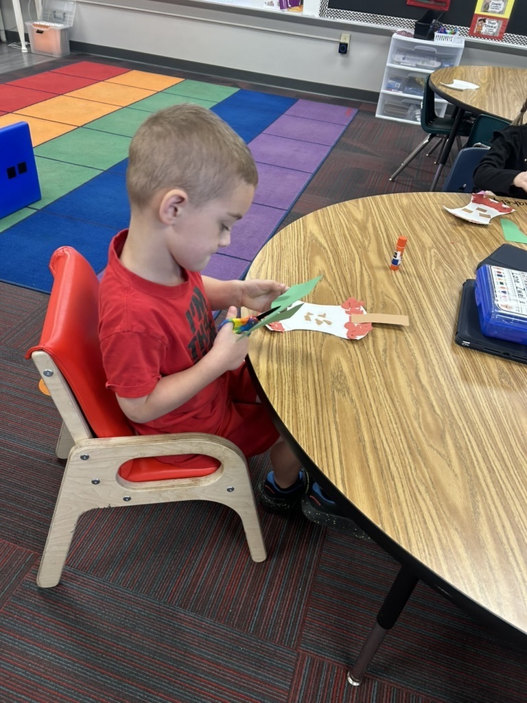 Boy sitting in a chair at a table working with scissors and a craft apple project.