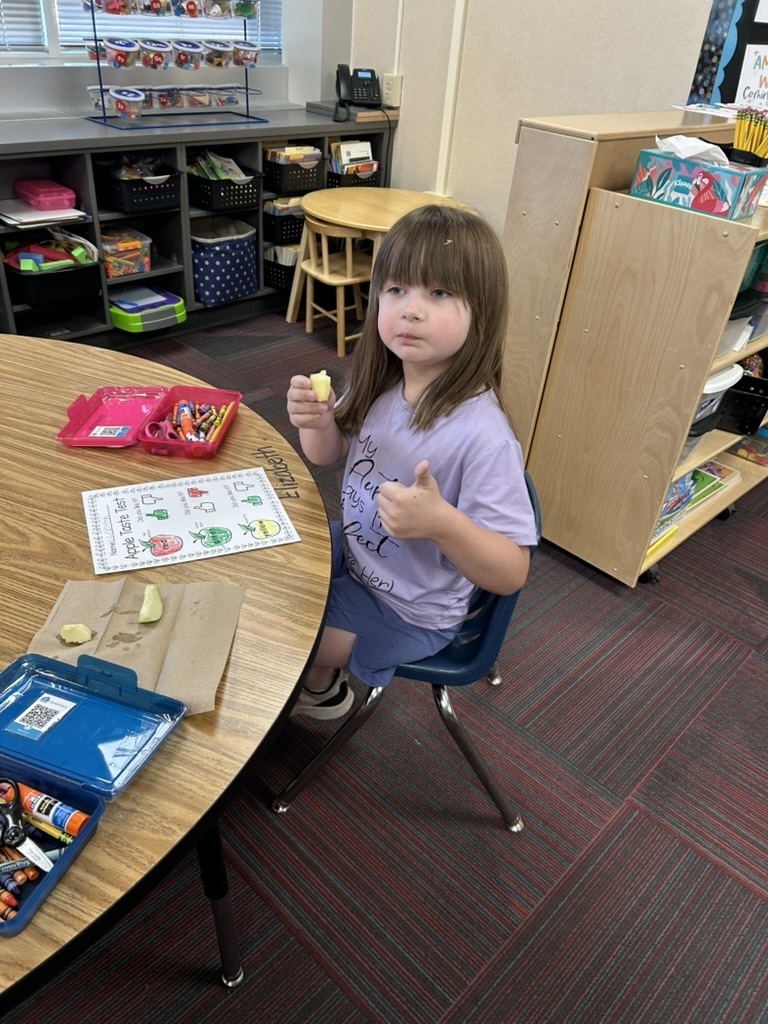 Girl sitting at a table with an apple in her hand with her thumb up and a paper on the table that says Apple Taste Test.
