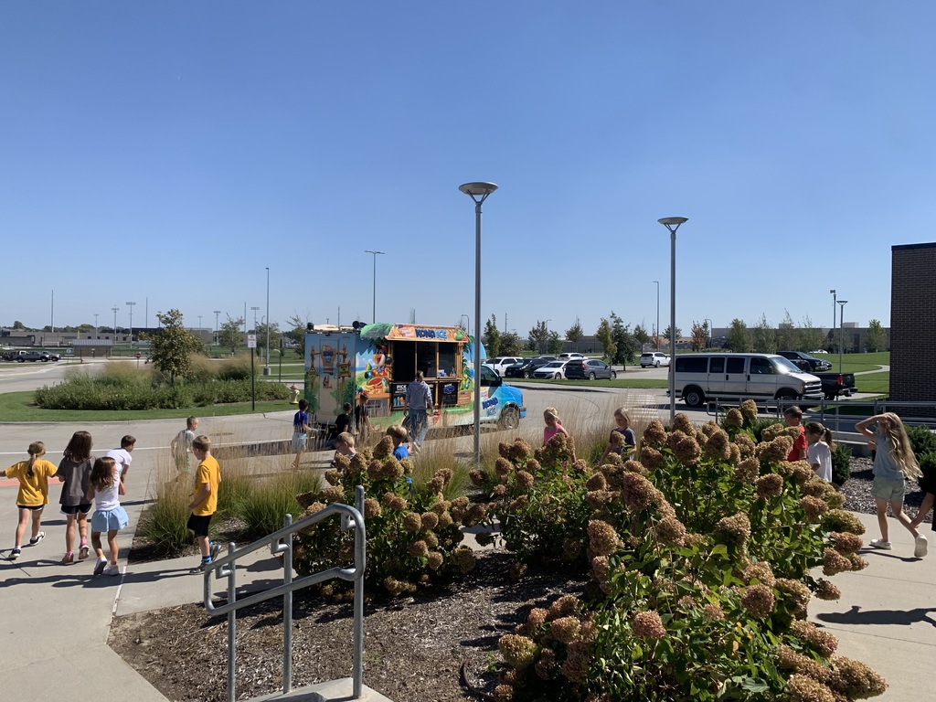 	A wider outdoor shot on a sunny day shows the Kona Ice truck parked in the distance with a group of children gathered around it, viewed from behind some decorative bushes.