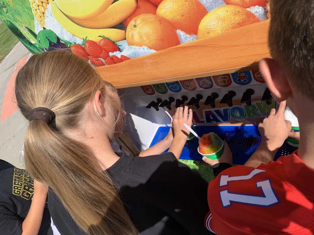 A close-up shot of a girl with her hair in a ponytail choosing her flavor from the self-serve Flavorwave of the Kona Ice truck, with her companion nearby. She is holding a colorful cup of shaved ice.