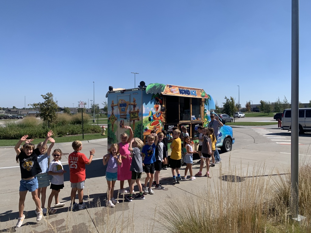 A long line of children, some waving excitedly, stand outdoors in front of a colorful Kona Ice shaved ice truck under a bright blue sky.