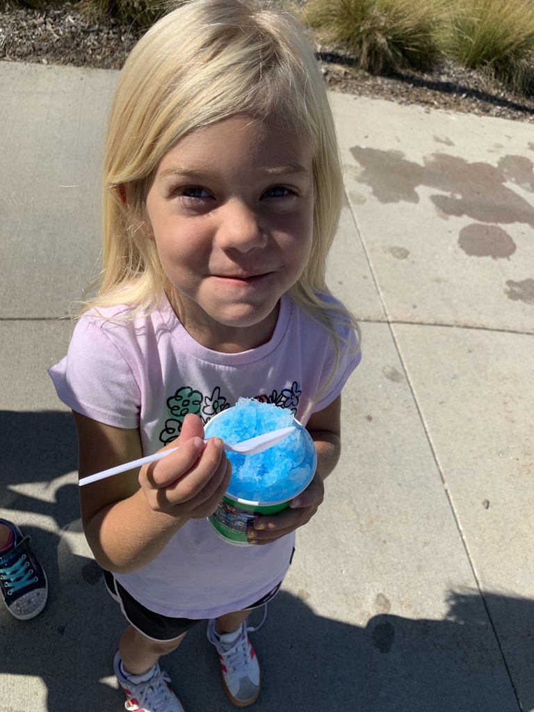 A young blonde girl in a pink shirt holds a cup of bright blue shaved ice (Kona Ice) and smiles at the camera on a sunny day.