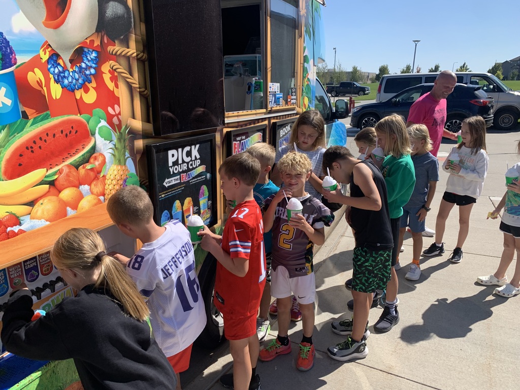 A close-up of several children lining up next to the Kona Ice truck's "Pick Your Cup" sign, with a few of them holding and drinking their cups of shaved ice.