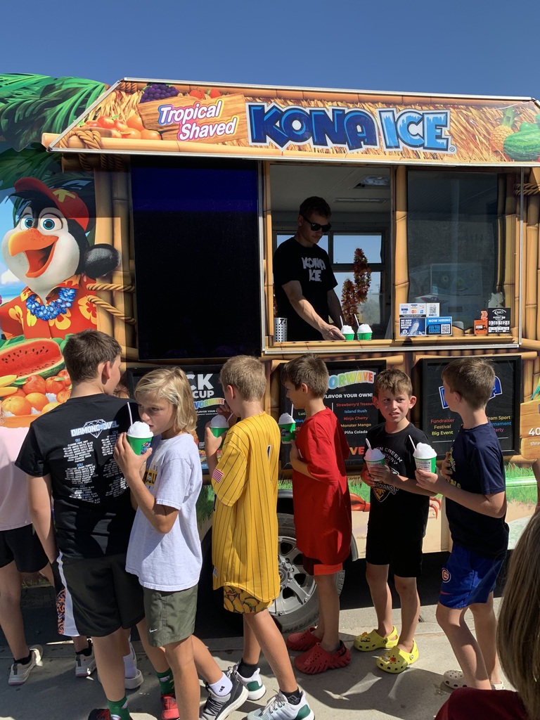	A group of boys stand in line next to the Kona Ice truck, talking and drinking from their cups. An adult employee in a black shirt is visible serving from the window.