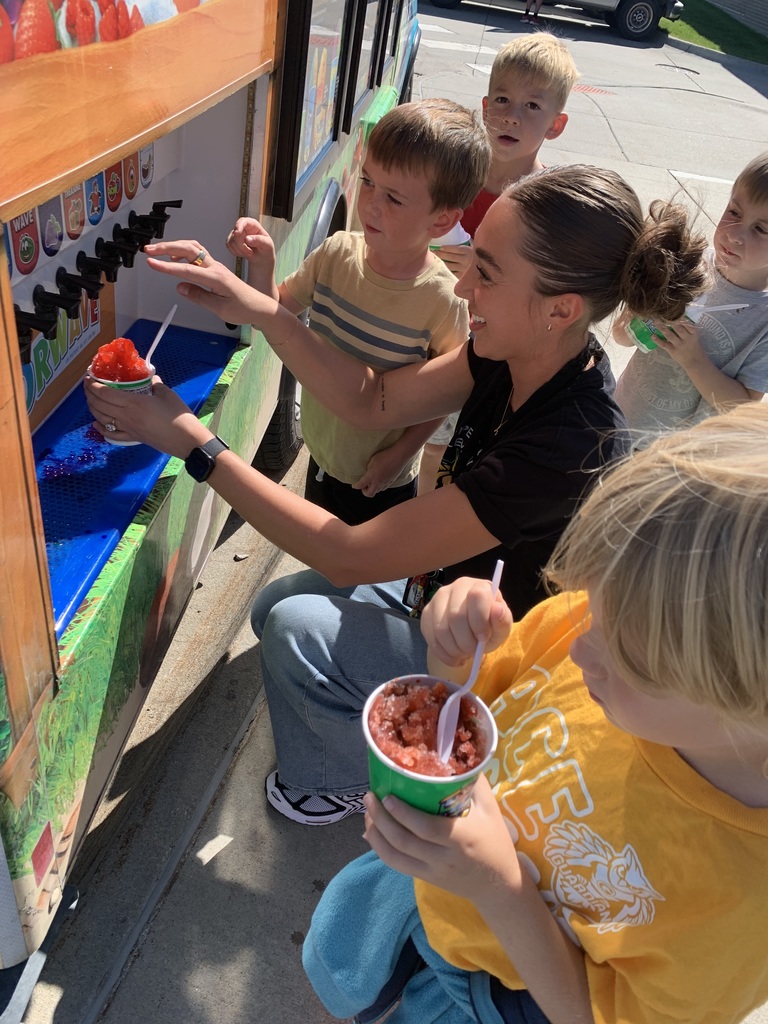 A woman kneels next to the Kona Ice Flavorwave to help a child fill their cup with red shaved ice flavor, as several other young children watch and wait.