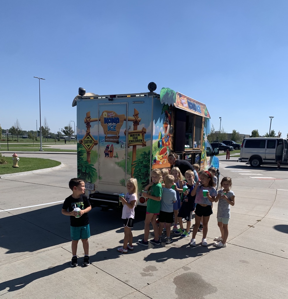 An outdoor photo on a bright, sunny day showing the colorful Kona Ice truck parked on an asphalt lot with a small group of children gathered around it.