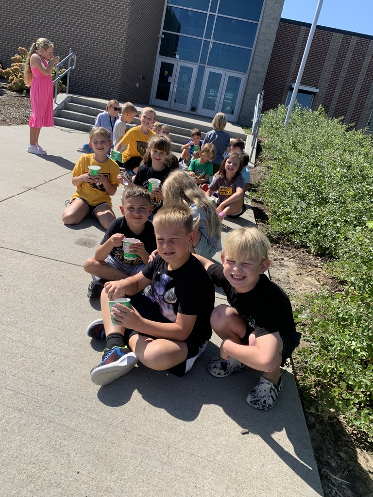 A group of smiling children, sitting on the sidewalk in front of a school building, happily holding their cups of shaved ice.