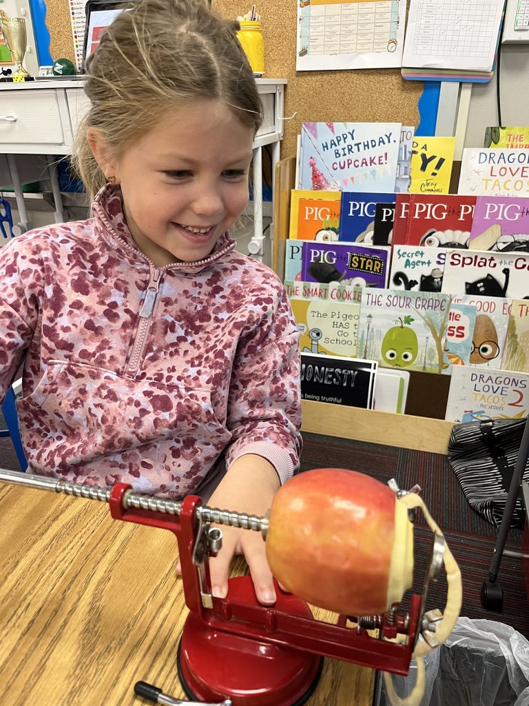 Girl with a pink jacket holding a handle to an apple peeler that is peeling a green apple.