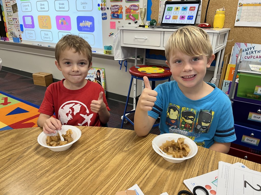 Two boys sitting at a table with their thumbs up with applesauce sitting on the table in white bowls.