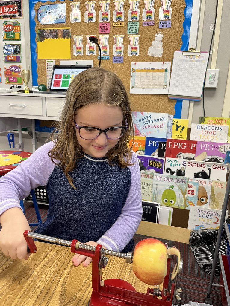Girl with a purple shirt holding a handle to an apple peeler that is peeling a red apple.