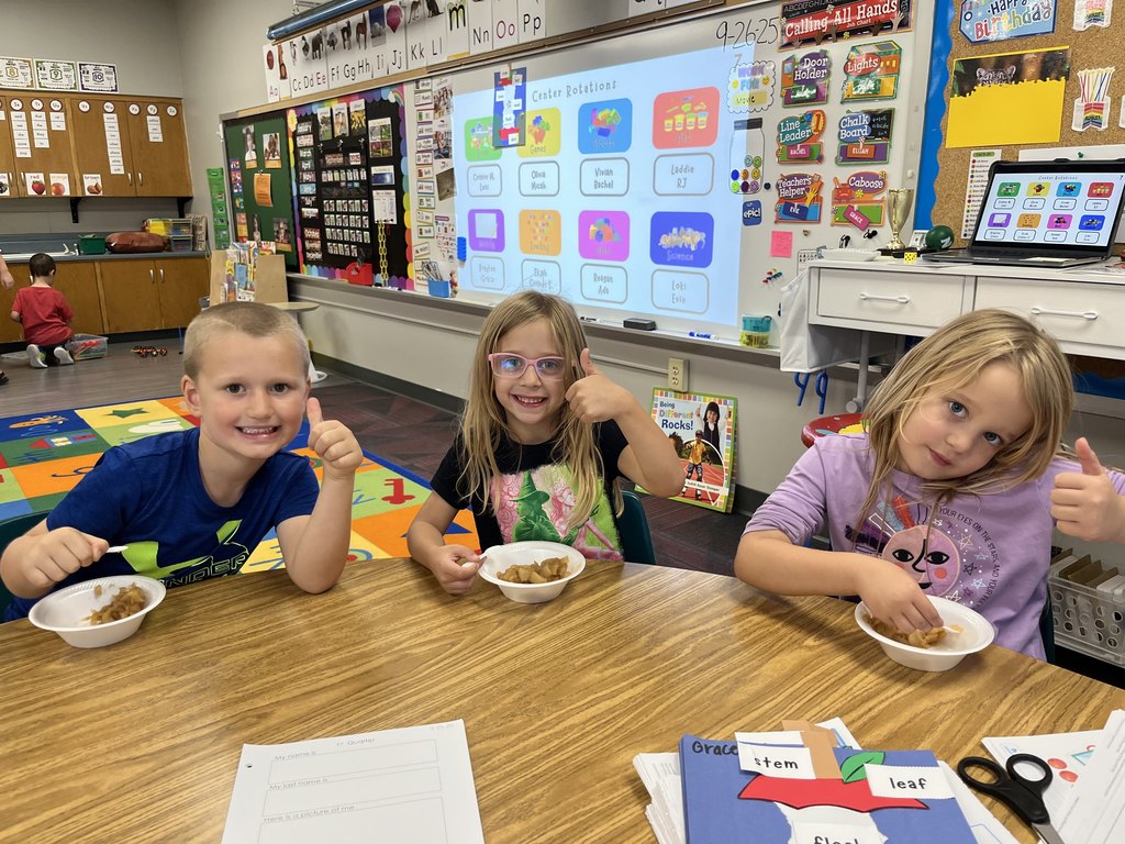 Three kids sitting at a table with their thumbs up with applesauce sitting on the table in white bowls.