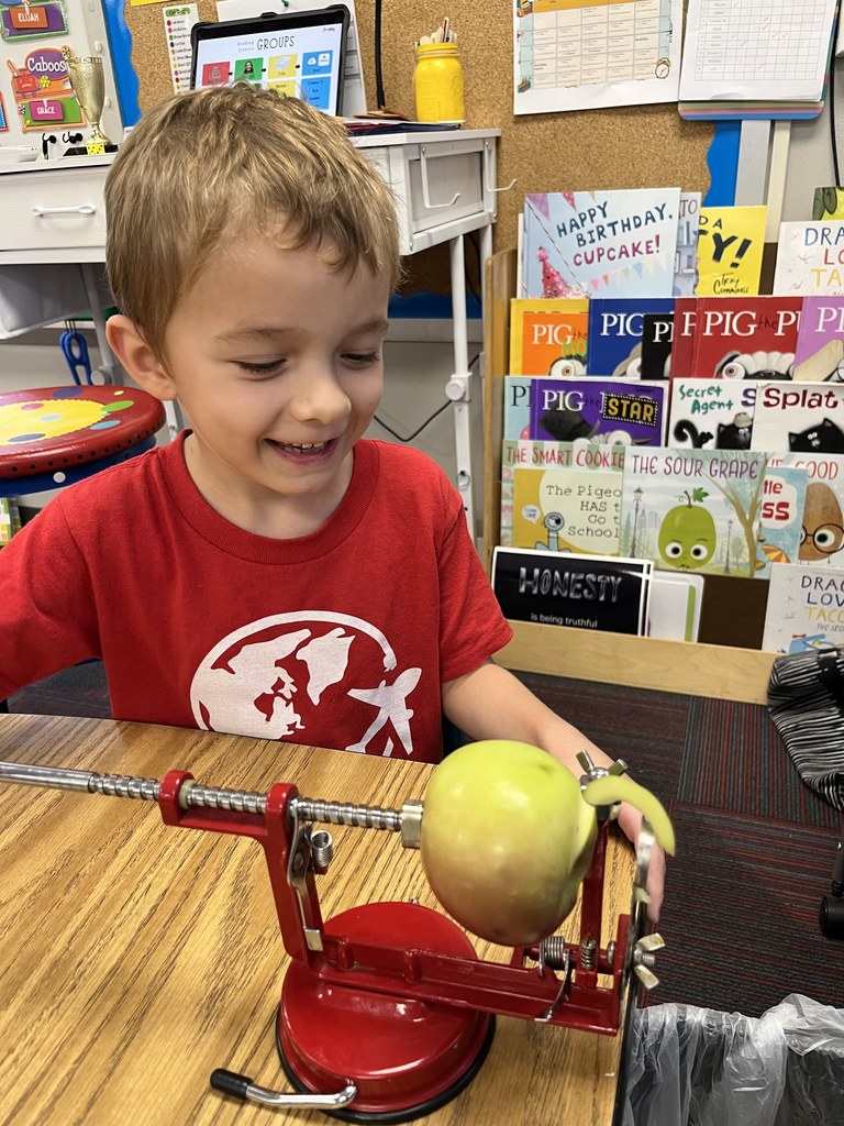 Boy with a red shirt holding a handle to an apple peeler that is peeling a green apple.