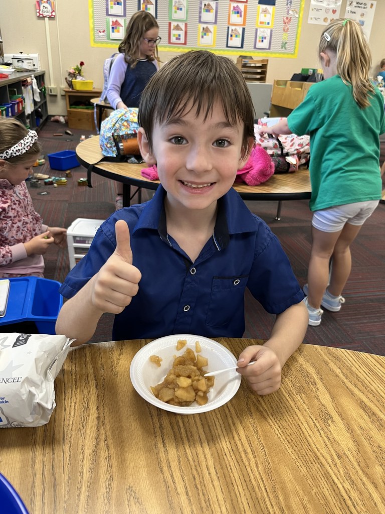 Boy with a blue shirt sitting at a table with his thumb up with applesauce sitting on the table in a white bowl.