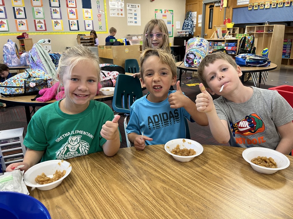Three kids sitting at a table with their thumbs up with applesauce sitting on the table in white bowls.