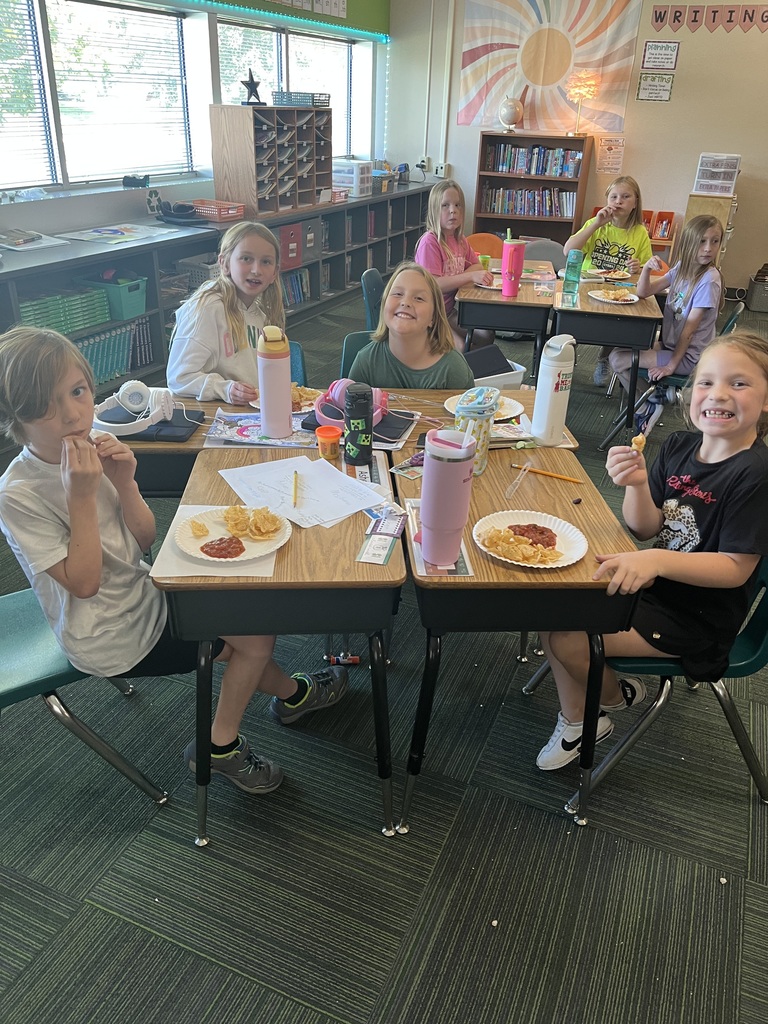 Group of students sitting in a classroom at desks with water bottles and plates filled with chips and salsa on the plates.