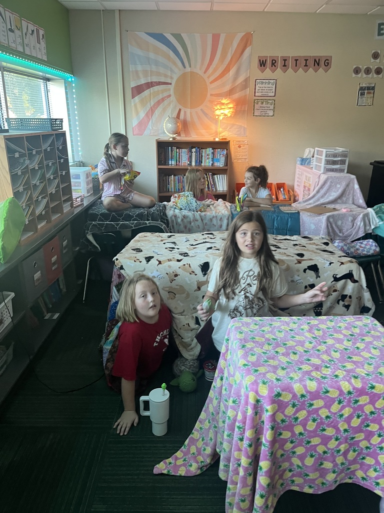 Group of students, some sitting, some laying in a room where blankets are covering desks and some students are laying on pillows.