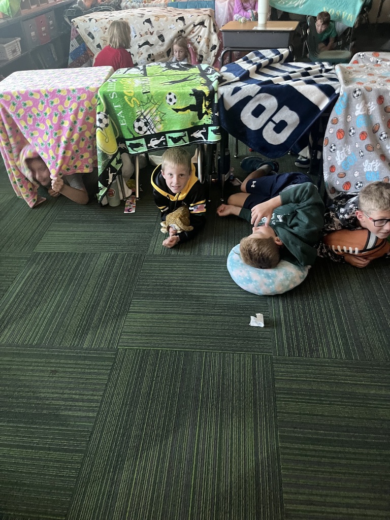 Group of students, some sitting, some laying in a room where blankets are covering desks and some students are laying on pillows.