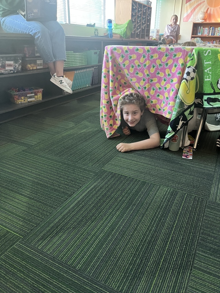Student laying on the ground smiling underneath a desk covered with a blanket.