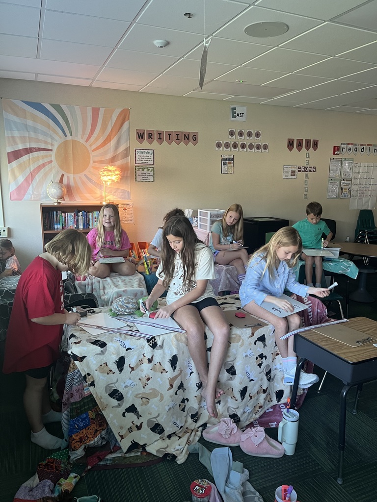Group of students, some sitting, some laying in a room where blankets are covering desks and some students are laying on pillows.