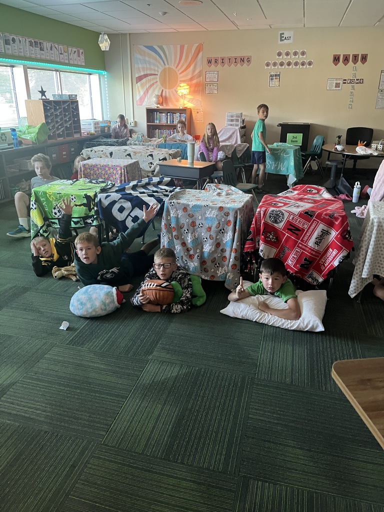 Group of students, some sitting, some laying in a room where blankets are covering desks and some students are laying on pillows.