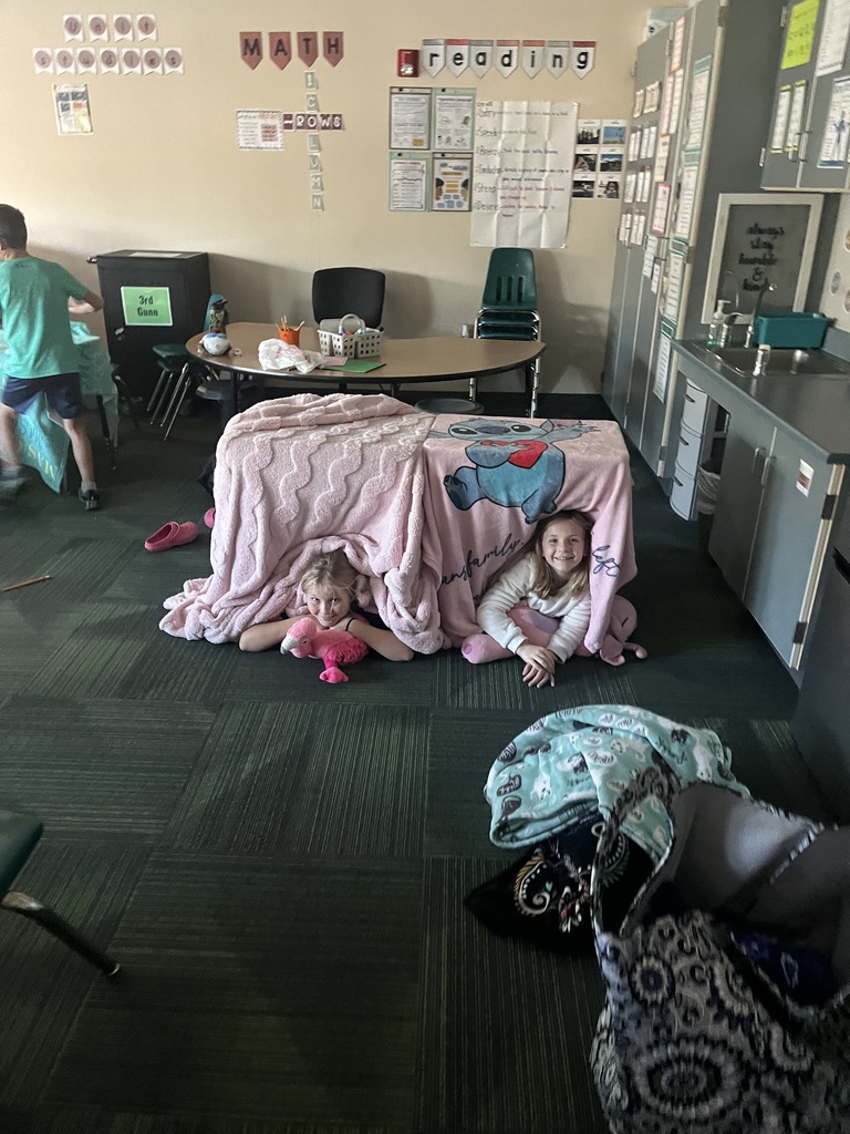Group of students, some sitting, some laying in a room where blankets are covering desks and some students are laying on pillows.
