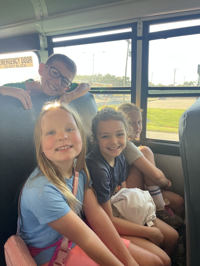 Three girls sitting on a bus in a seat while smiling and a boy standing holding onto the edge of the seat smiling.
