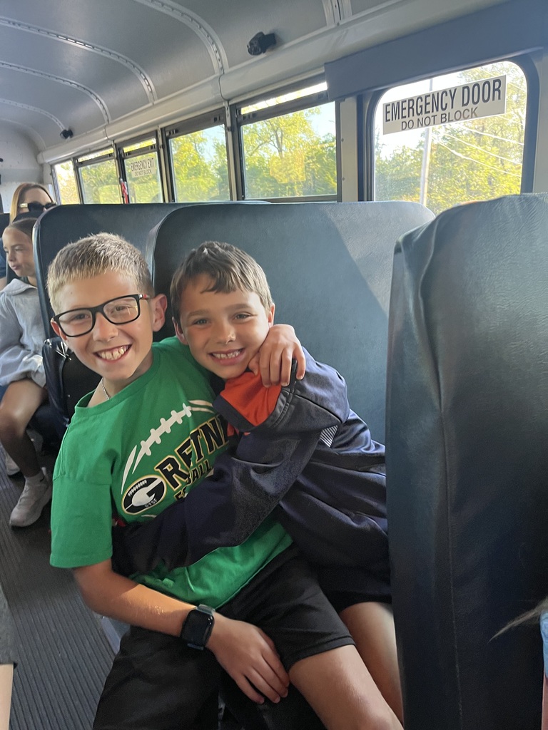 Two boys sitting on a bus in a seat while smiling and giving each other a hug.