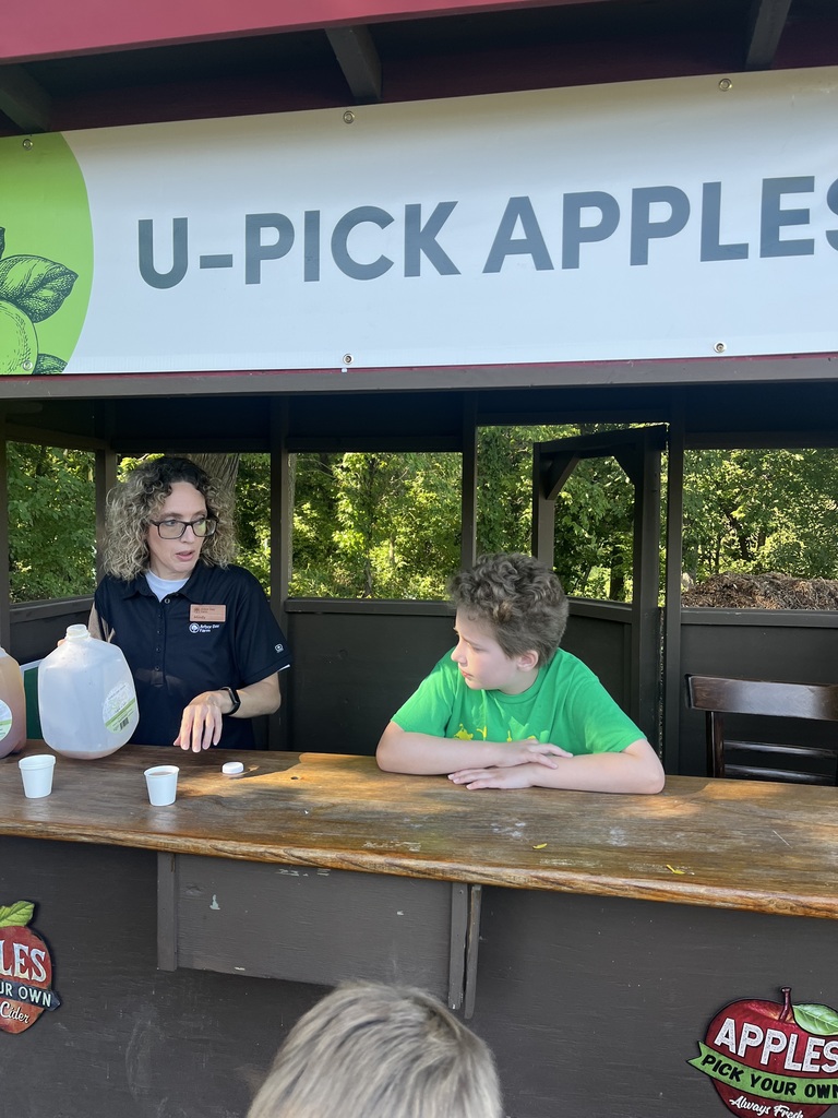 Boy stands behind a table with an adult holding a jug of apple cider.