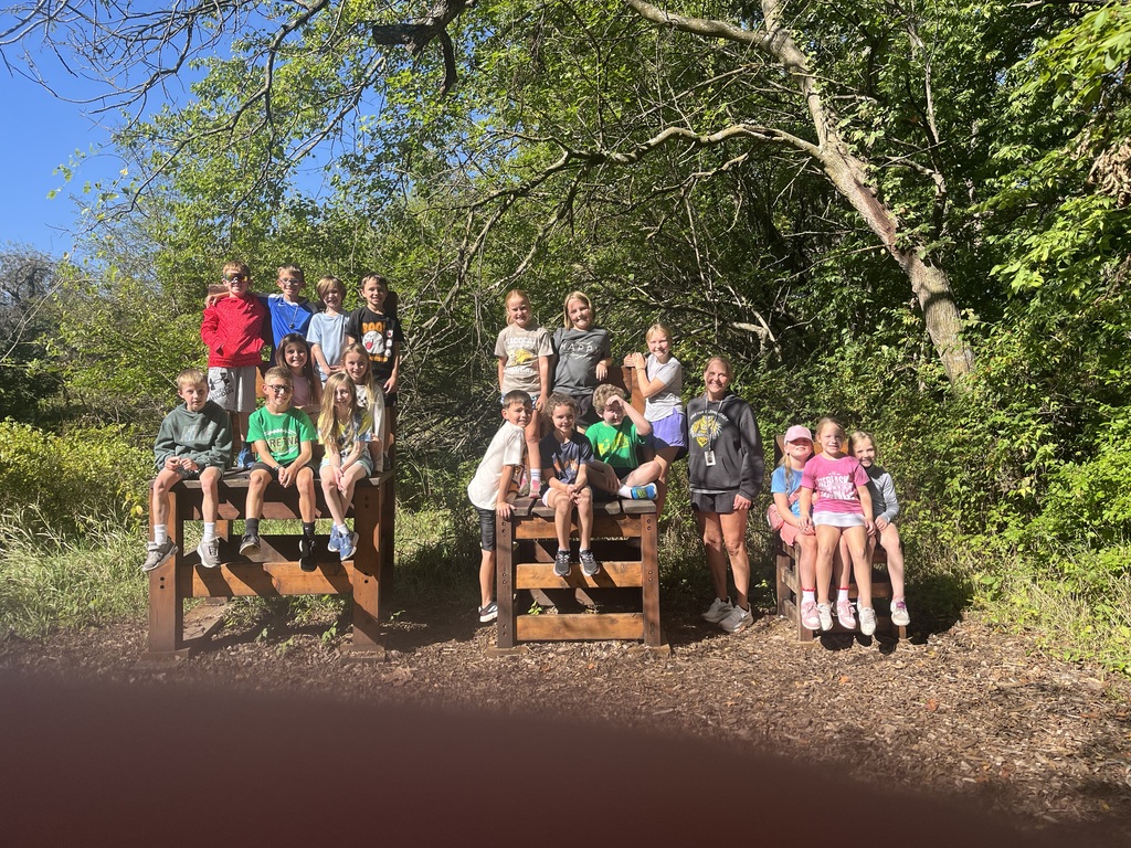 Group of students, some sitting and some standing on giant chairs with trees in the background.