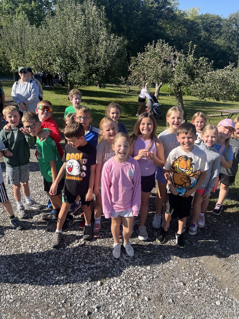 Group of third grade students standing on a gravel road smiling with apple trees in the background.