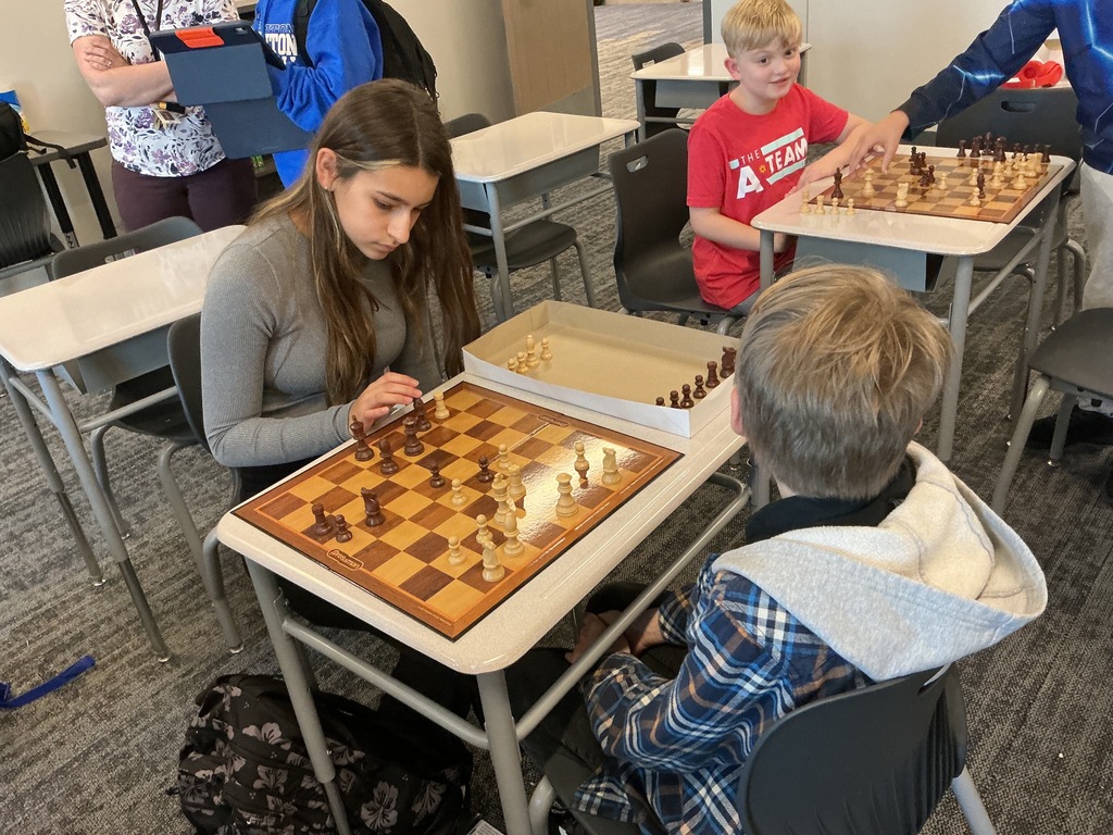Photo of two students playing chess at a desk while a nearby student watches.