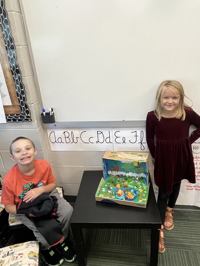 Boy and girl smiling standing next to a brown shoe box with a diorama of an animal made with art supplies inside.