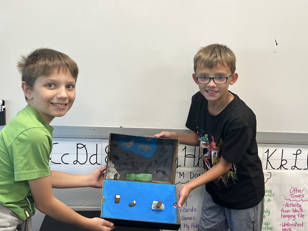Two boys smiling holding a blue shoe box with a diorama of an animal made with art supplies inside.