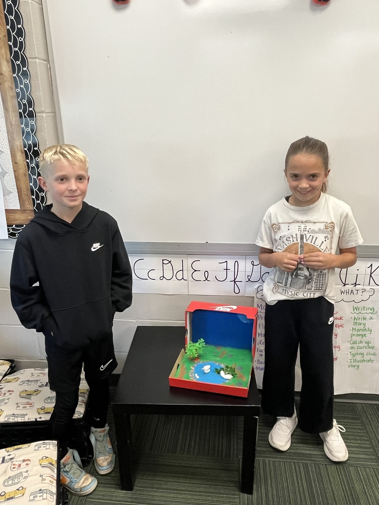 Boy and girl smiling standing next to a red shoe box with a diorama of an animal made with art supplies inside.