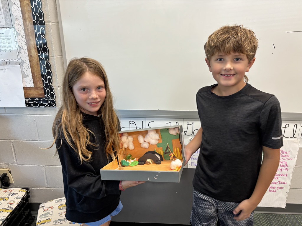 Boy and girl smiling holding a gray shoe box with a diorama of an animal made with art supplies inside.