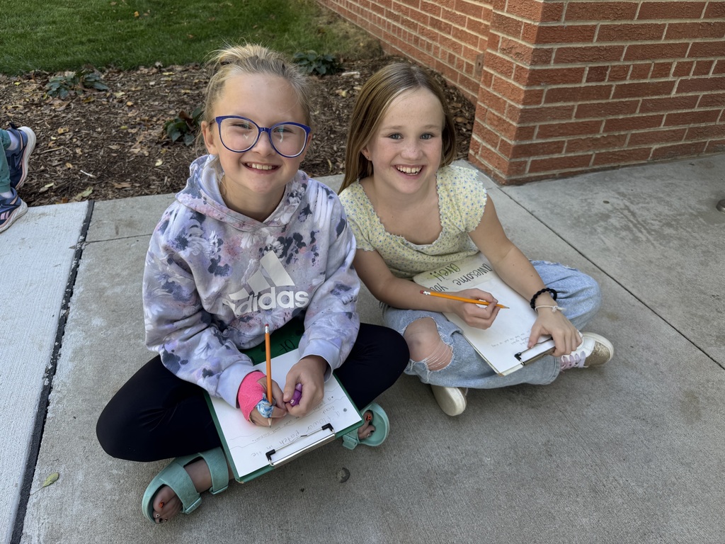 Two girls sitting outside on cement with clipboards and papers in their hands.