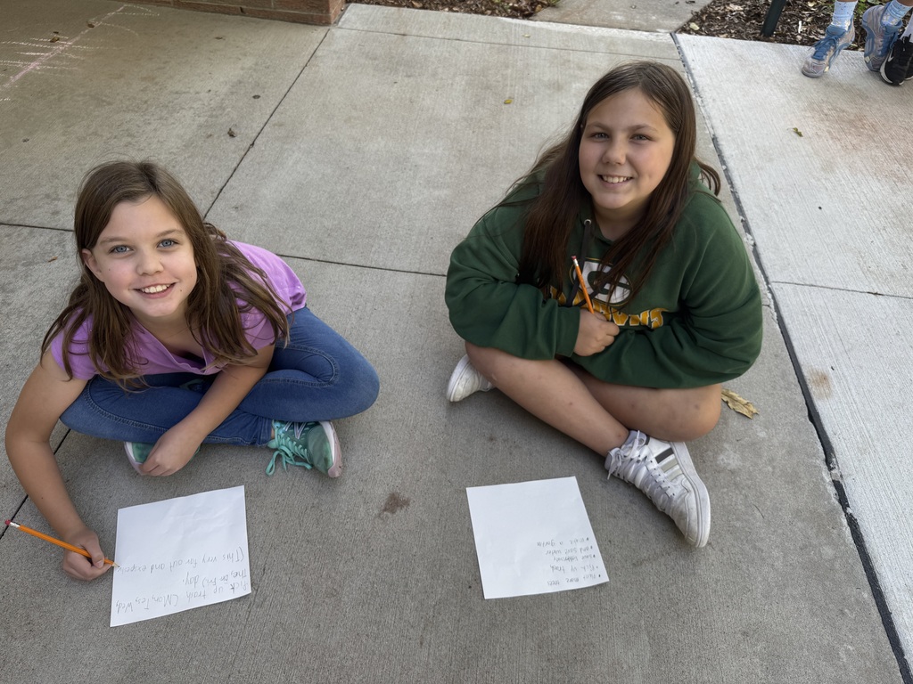 Two girls sitting outside on cement with clipboards and papers in their hands.
