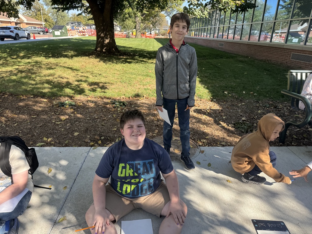 Two boys sitting and standing outside on cement with clipboards and papers in their hands.